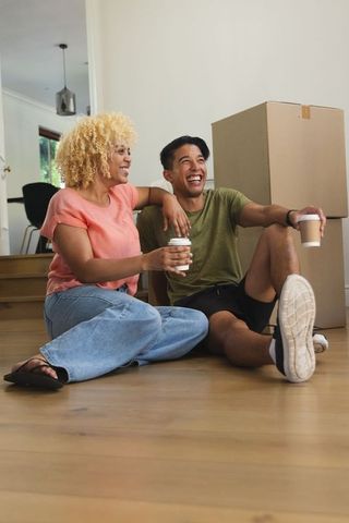 Couple sitting on hardwood floor laughing and holding takeaway coffee near moving boxes