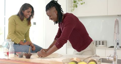 Mixed-race couple kneading dough in sunlit modern kitchen, woman smiling on countertop