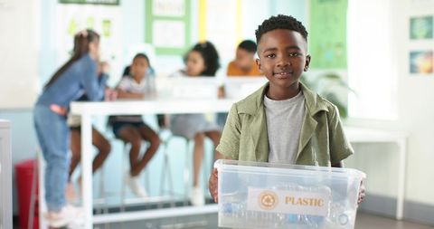 African american boy holding plastic recycling bin in classroom for youth sustainability
