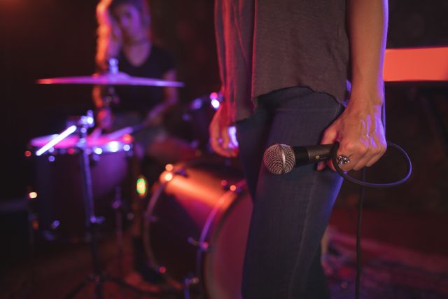 Female singer holding microphone while drummer performs in illuminated nightclub. Ideal for use in articles about live music, nightlife, entertainment, and band performances. Suitable for promoting concerts, music events, and nightclubs.