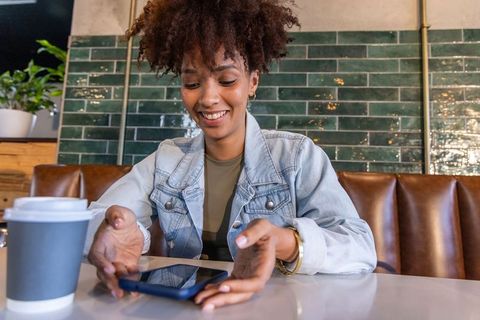 African woman smiling reaching for smartphone at cafe with coffee cup and denim jacket
