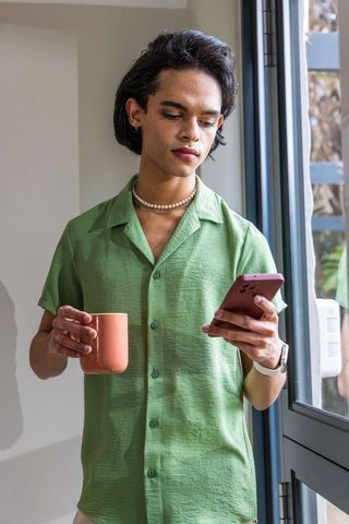 Calm asian man checking phone while holding coffee by sunlit glass door at home