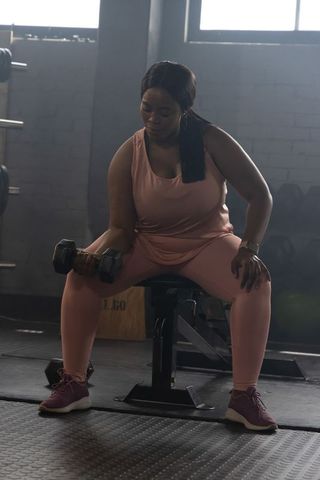 African american woman performing seated dumbbell curl on bench wearing pink activewear