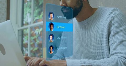 Mid-adult man typing on laptop while interacting with holographic contact list UI at home