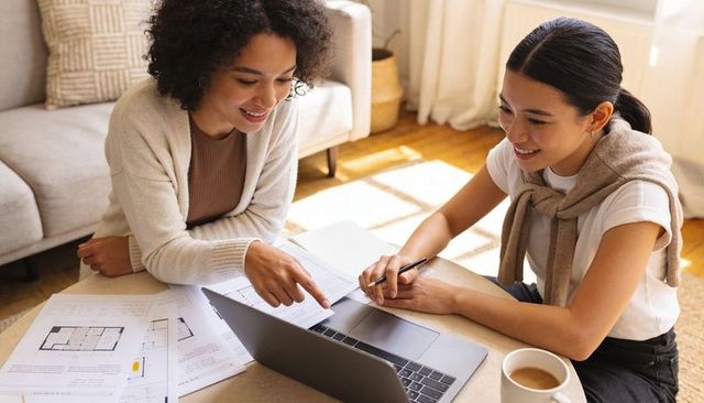 Two women collaborating at home on laptop and floor plans planning renovation project