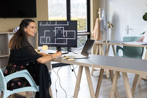 Indian architect reviewing floor plan in modern design studio with laptop and monitor