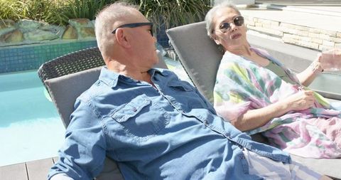 Senior asian couple reclining and chatting on poolside chaise lounges in sunny backyard