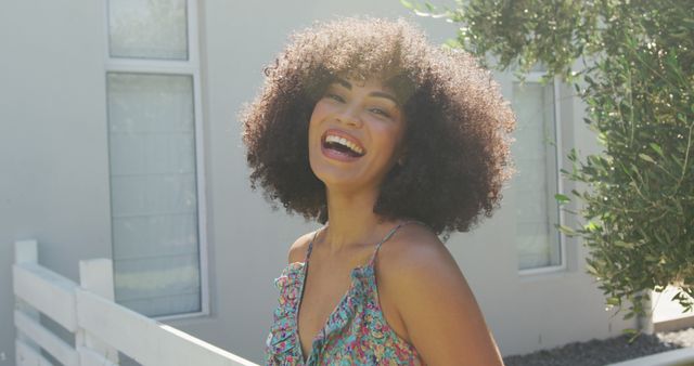 Young woman with curly hair is smiling brightly while standing outside on a sunny day. Natural light highlights her cheerful expression. She is wearing a casual summer dress, exuding joy and radiance. Ideal for promoting positive emotions, outdoor activities, summer fashion, and carefree lifestyle content.