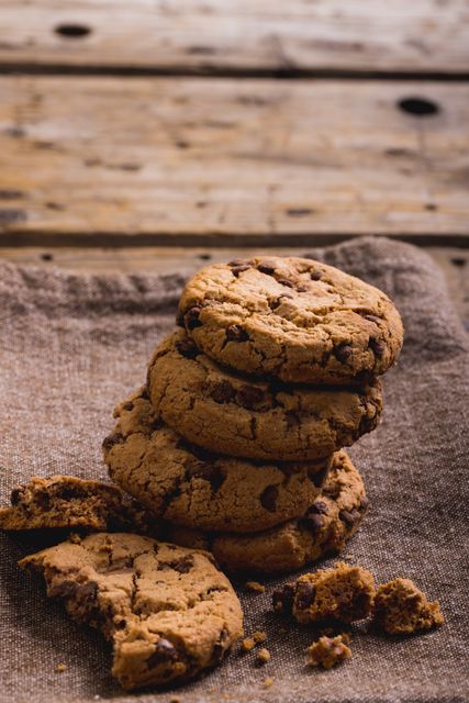 Close-up of a stack of chocolate chip cookies on burlap at a wooden table. Crumbs are scattered around, adding a rustic and homemade feel. Ideal for use in food blogs, recipe websites, advertisements for bakeries, or social media posts about baking and desserts.