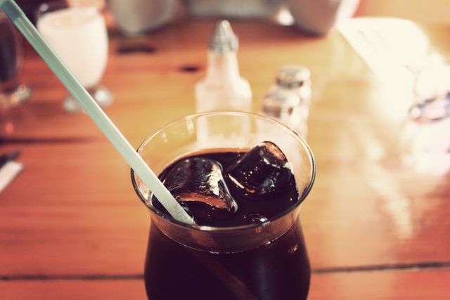 Close-up of an iced coffee in a glass with ice cubes and a straw, placed on a wooden table. Ideal for use in hospitality, food and beverage marketing, summer promotions, or café atmosphere themes.