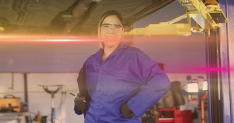 Smiling female mechanic inspecting vehicle under lift with clipboard and protective gear