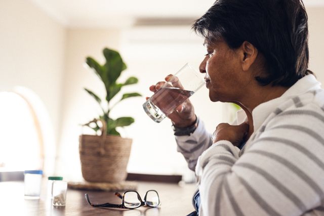 Side view of biracial mature woman with short hair drinking water while eating medicines at home. Eyeglasses, unaltered, healthcare, pills, sickness, treatment and retirement concept.