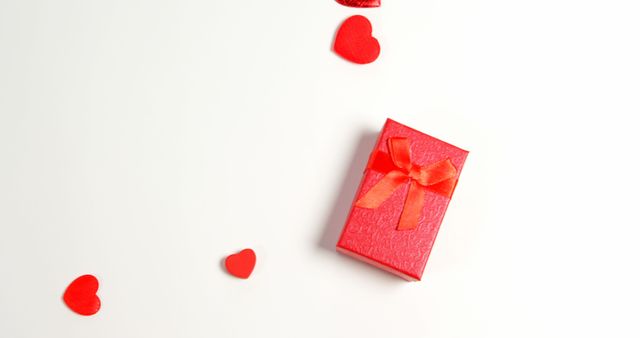 A red gift box tied with a ribbon is surrounded by small red hearts on a white background, with copy space. It suggests a romantic occasion like Valentine's Day or an expression of love and affection through gift-giving.