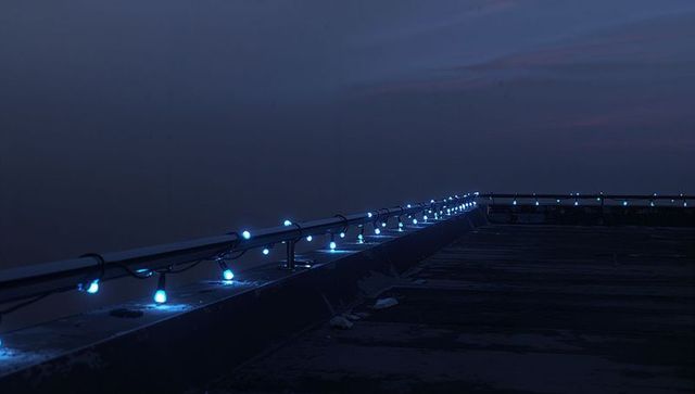Rooftop railing glowing with blue string lights leading into urban twilight horizon