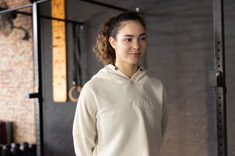 Young woman standing in industrial gym wearing light hoodie beside rig and kettlebells