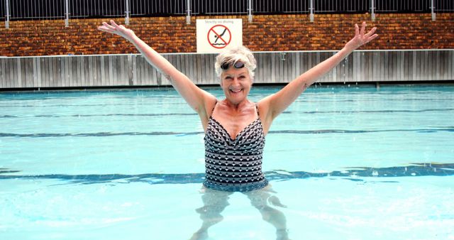 Senior woman standing in clear, outdoor swimming pool, smiling and lifting hands above head. Ideal for projects on senior health, active lifestyle, summer activities, leisure, and well-being.