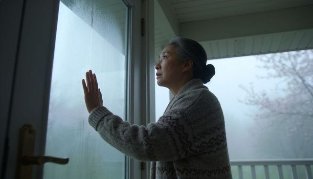 Asian woman pressing hand to fogged glass door, gazing into misty morning on porch