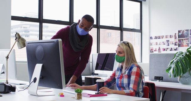 Office colleagues working together on a project in a modern office setting, both wearing face masks. The image depicts a collaborative working environment with attention to pandemic safety measures. Useful for illustrating workplace safety, teamwork, modern office culture, and professional communication.