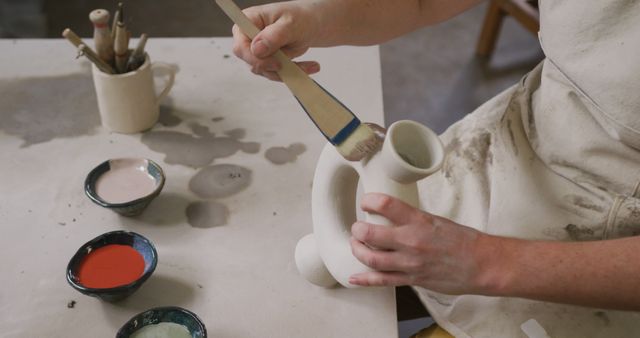 Mid section of female potter wearing apron using glaze brush to paint on pot at pottery studio. hygiene and social distancing in the pottery studio during coronavirus covid 19 pandemic.