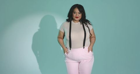 Mid-30s woman standing in beige top with suspenders and pastel pink trousers on teal backdrop