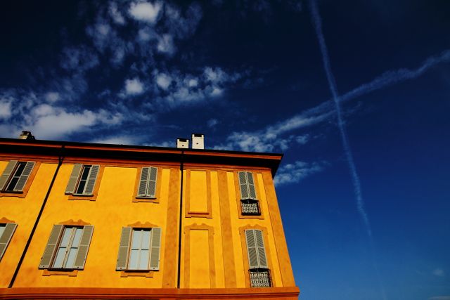 Yellow building with closed window shutters is pictured against a clear blue sky with scattered clouds and a visible airplane contrail. Suitable for illustrating urban scenes, European cities, architectural details, tourism, and travel brochures.