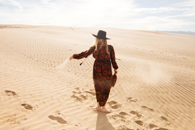 Woman walking barefoot through expansive desert sand dunes wearing a flowing dress and wide-brimmed hat. Serene and adventurous atmosphere. Ideal for travel blogs, fashion editorials, lifestyle and mindfulness campaigns, and social media content focused on exploration and tranquility.