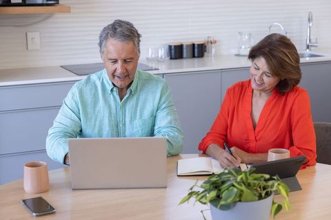 Mature couple working remotely at kitchen table, using laptop and tablet for planning