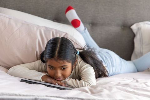 Asian girl lying on bed watching tablet on pastel pink bedding, cozy bedroom scene