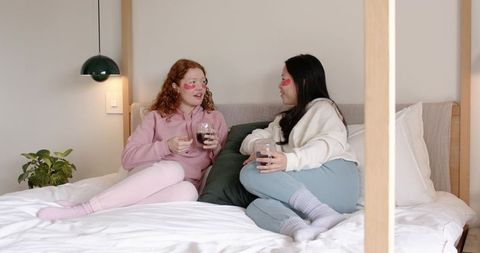 Two women reclining on bed wearing pastel loungewear and under-eye patches holding wine