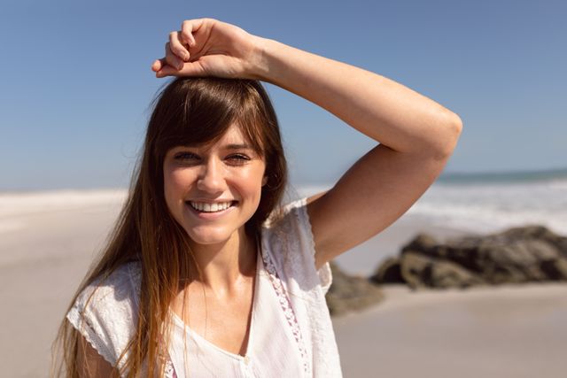 Young woman smiling and enjoying a sunny day at the beach. Ideal for use in travel brochures, lifestyle blogs, vacation advertisements, and wellness promotions. Perfect for conveying themes of happiness, relaxation, and carefree living.