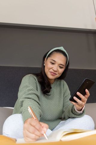 Asian woman journaling on bed while checking phone in cozy minimalist bedroom