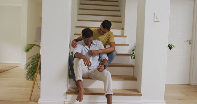 Smiling biracial gay male couple sitting on stairs holding hands embracing and talking. staying at home in isolation during quarantine lockdown.