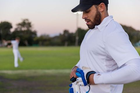 Male cricketer adjusting batting gloves at dusk on outdoor practice field under floodlights