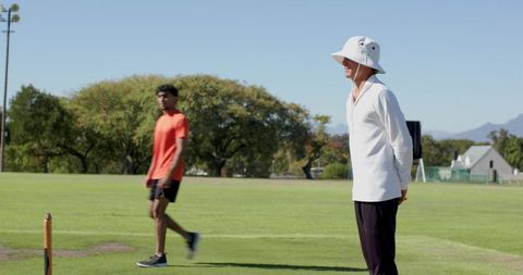 Cricket umpire standing on sunlit pitch wearing bucket hat observing players on field