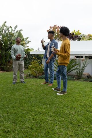 Diverse teens enjoying backyard hangout with football and bottled drinks, suburban friends
