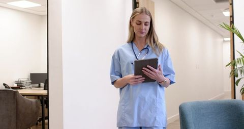 Female nurse checking tablet while walking clinic corridor wearing scrubs and stethoscope