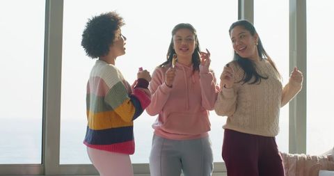 Three women singing and swaying by ocean window holding microphones during cozy jam