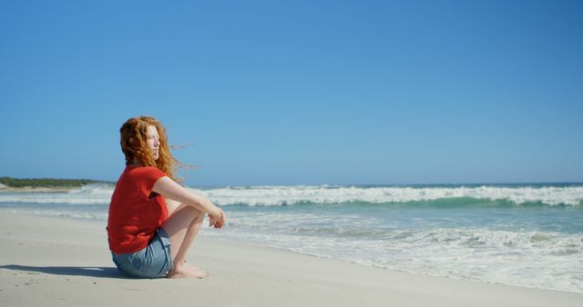 Young woman sitting on a sandy beach with the ocean spreading out before her, under clear blue skies. Perfect for concepts of relaxation, tranquility, leisure, and summer vacations. Suitable for travel brochures, wellness blogs, lifestyle articles, and therapeutic content.