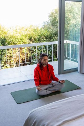 African american man meditating on green yoga mat in sunlit bedroom with balcony plants