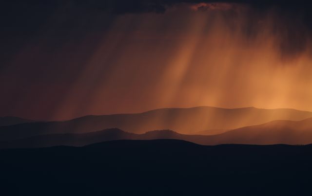 Golden sunbeams breaking through dark clouds over a mountain range at sunset. This dramatic and scenic image can be used for travel websites, nature documentaries, desktop wallpapers, inspirational backgrounds, and artistic projects highlighting the beauty of nature.