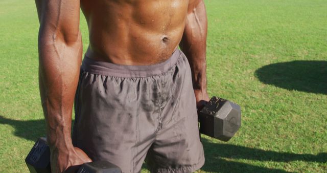 Tired, shirtless african american man dropping dumbbells and recovering after exercising outdoors. cross training for fitness at a sports field.