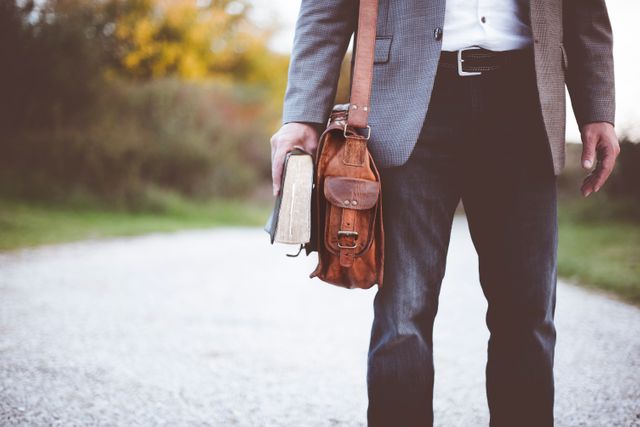 Man in business casual attire holding a book and leather bag while standing on a gravel path outdoors. Image can be used for themes related to education, leisure activities, outdoor adventures, business travel, and autumn settings.