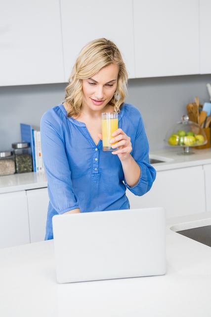 Blonde woman holding a glass of juice while using a laptop in a modern kitchen, suggests multitasking and a healthy lifestyle. Ideal for use in advertisements or articles about work-from-home setups, healthy living, or modern kitchen designs.