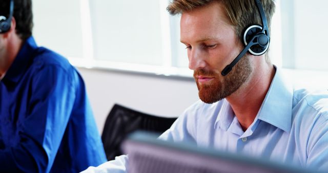 A Caucasian man wearing a headset is focused on his computer screen in an office setting, with copy space. His professional demeanor suggests he may be engaged in customer service or a similar support role.