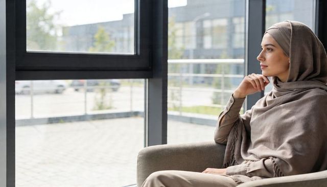 Contemplating woman wearing neutral hijab gazing out window from armchair in modern lounge
