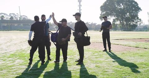 Sunlit amateur baseball team high-fiving on morning field with gloves and duffels