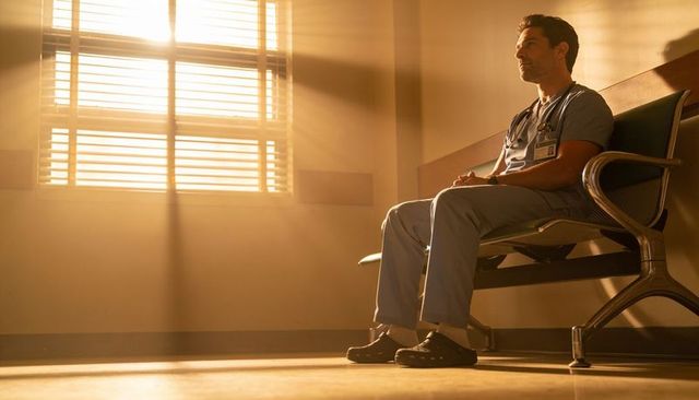 Thoughtful clinician sitting in sunlit hospital corridor reflecting after long shift
