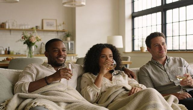 Friends relaxing on couch sharing wine and cozy blanket in sunlit open-plan living room