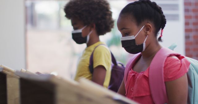 Image shows school children wearing face masks and backpacks as they study in a classroom. They appear focused and engaged. This type of image can be used for educational articles, pandemic-related educational material, back-to-school promotions, and public health campaigns.