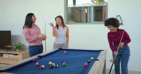 Diverse female friends playing pool laughing around blue-felt billiards table game night